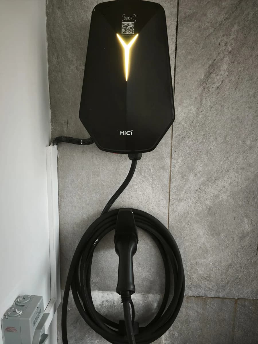 A black HiCi charger with its Y-shaped light lit, on a grey stone-tiled wall, the cable coiled below.