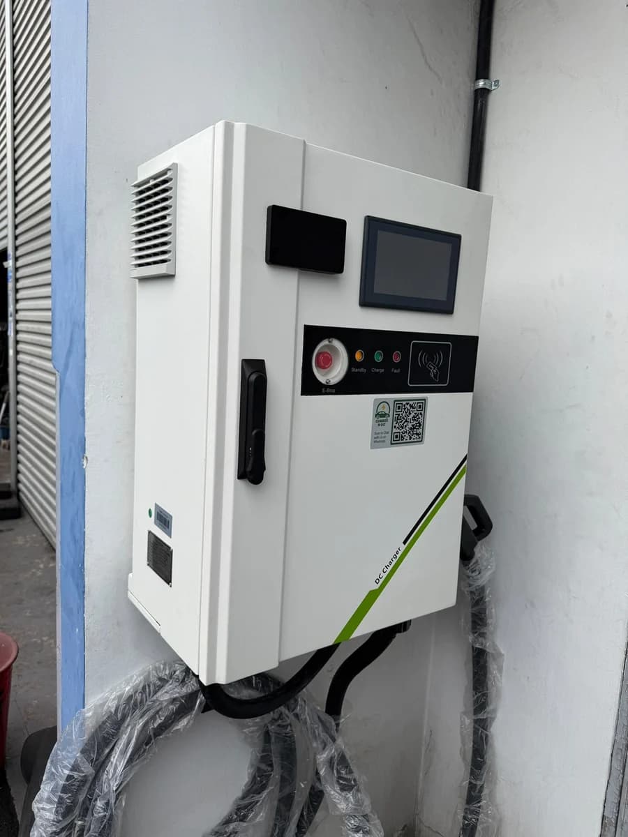 A white wall-mounted DC fast charger with a touchscreen and emergency stop, beside a roller shutter.