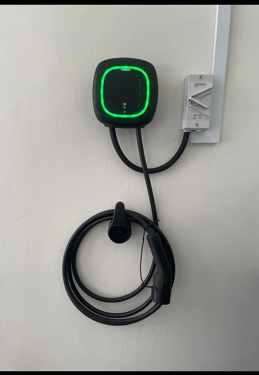 A dark wall charger with its status ring lit bright green, a white isolator beside it and the cable coiled on a hook below.