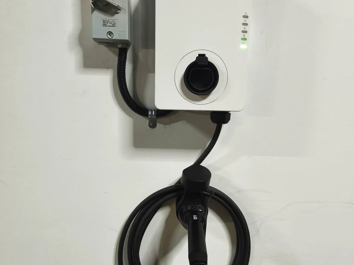 A white socketed EV charger with a black socket cap and an LED status strip, an isolator box above to the left and a thick supply cable entering through a gland underneath.
