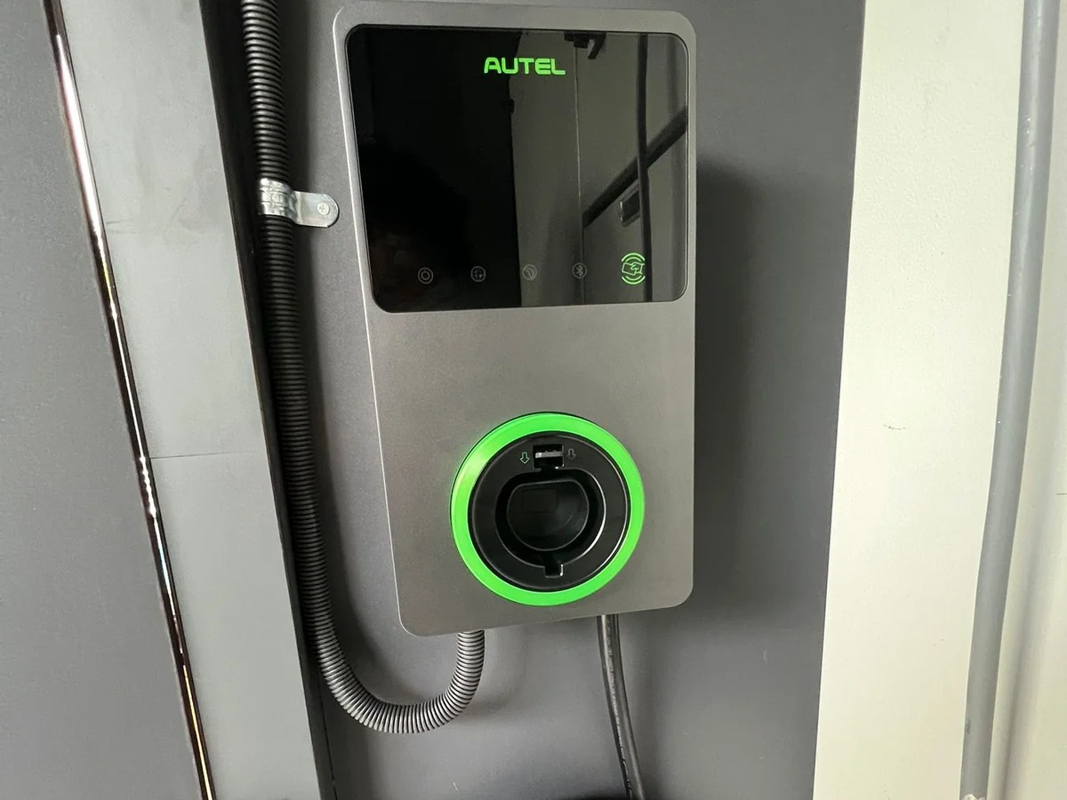 A grey Autel wall charger with a black glass display panel and a green-lit socket ring, mounted on a dark backing panel with flexible conduit running into it from the left.