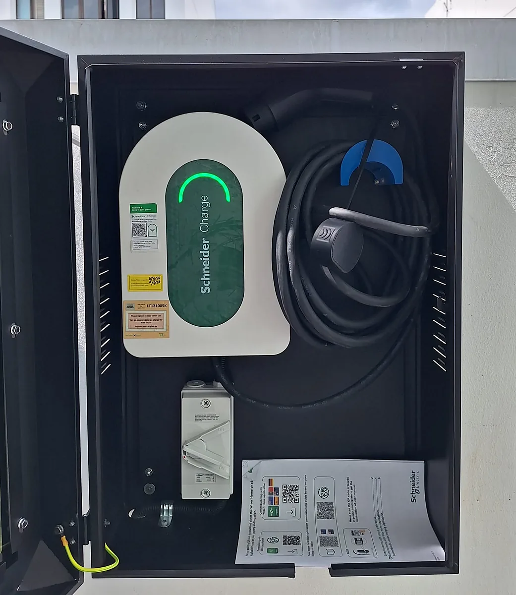 A wall-mounted EV charger inside a steel enclosure with the glazed door swung open, the isolator below it, the cable coiled beside the unit and the commissioning documents resting on the cabinet floor.