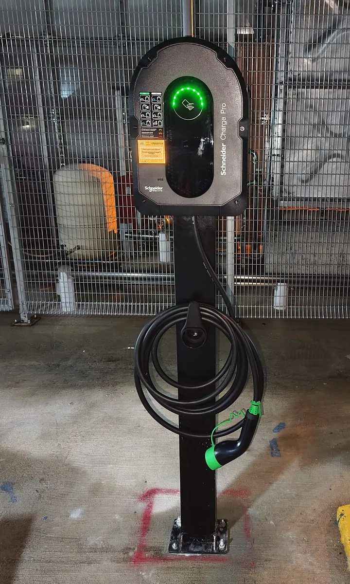 An EV charger on a black standalone steel post, shown from the charger head down to the base plate bolted to the concrete, its cable coiled around the post and the connector holstered.