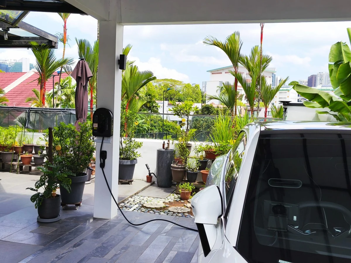 A black AC wall charger on a pillar in a covered landed-home carport, its cable running to a white car parked alongside, with potted plants and palms around the terrace