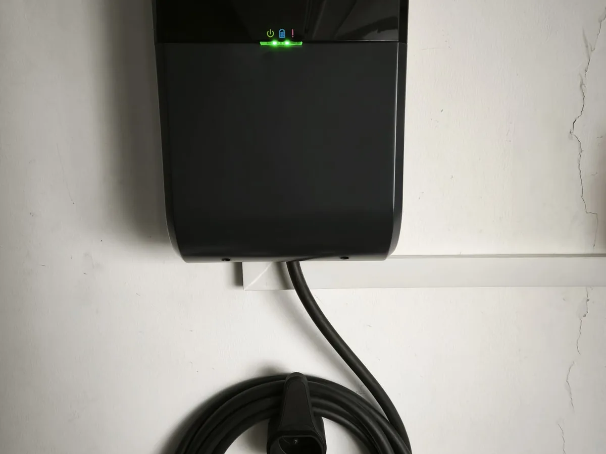 A black wall charger with a green LED status bar near the top, mounted on a white wall with its cable coiled beneath.