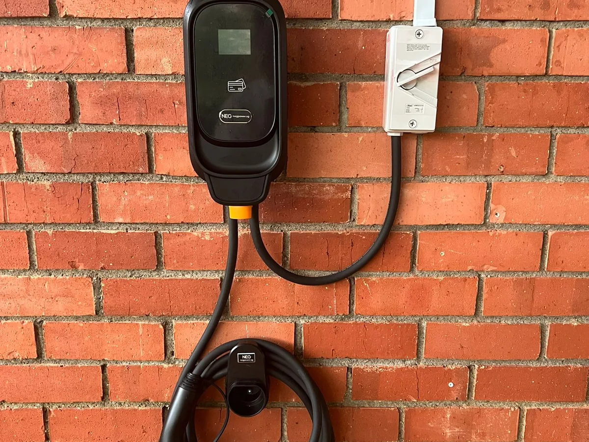 A black NEG wall charger on a red brick wall with a white isolator switch beside it and the tethered connector hanging below.