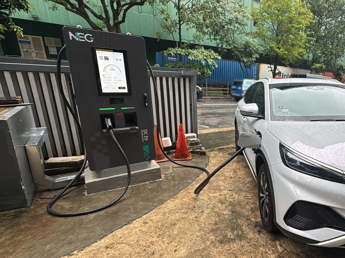 A NEG DC fast charger with twin cables on a concrete apron, a white SUV plugged in beside it and stacked shipping containers in the background.