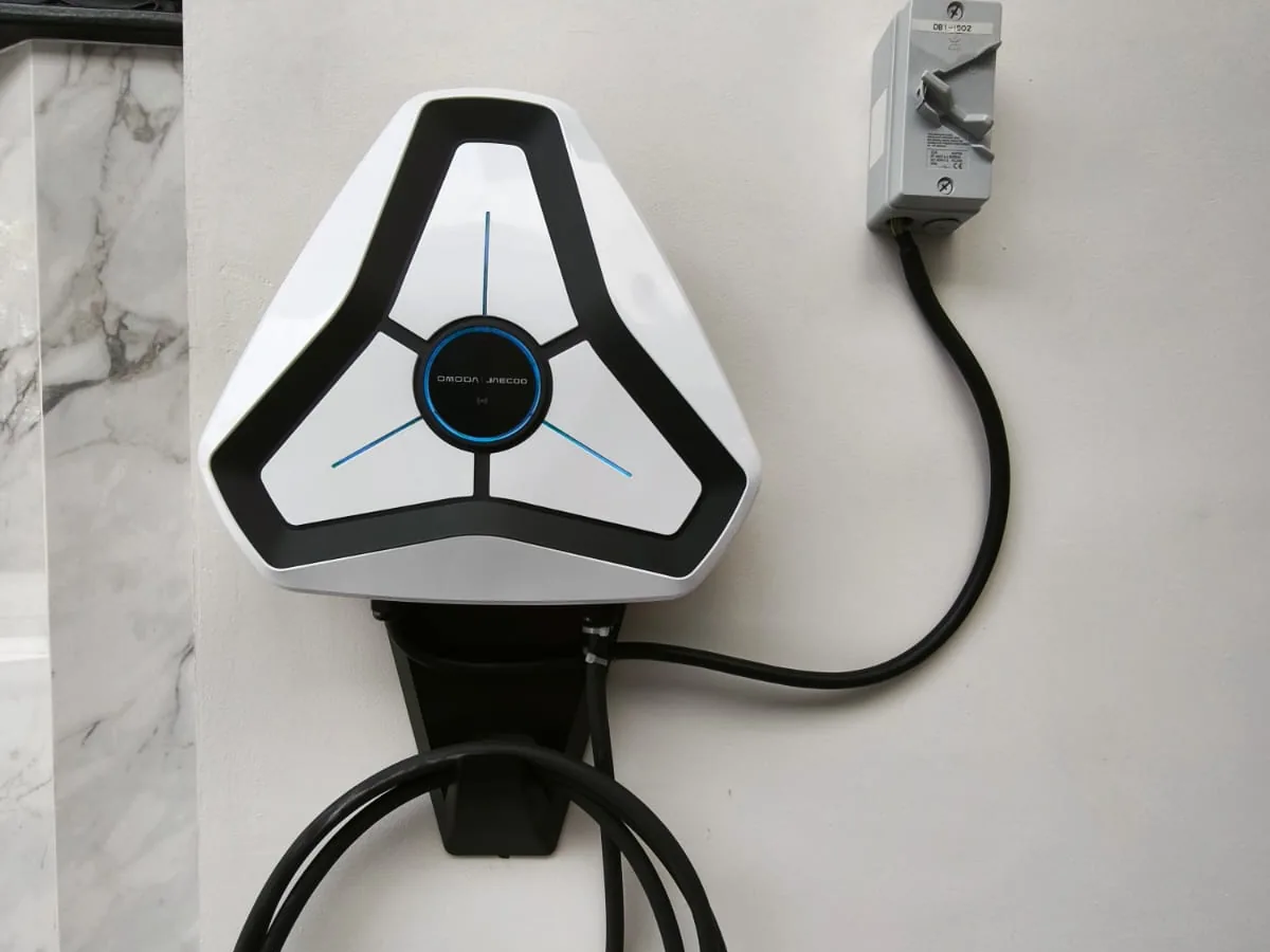 A white three-spoke wall charger with blue illuminated accents and an OMODA and JAECOO badge on its central hub, mounted on a marble-effect wall with an isolator switch beside it.