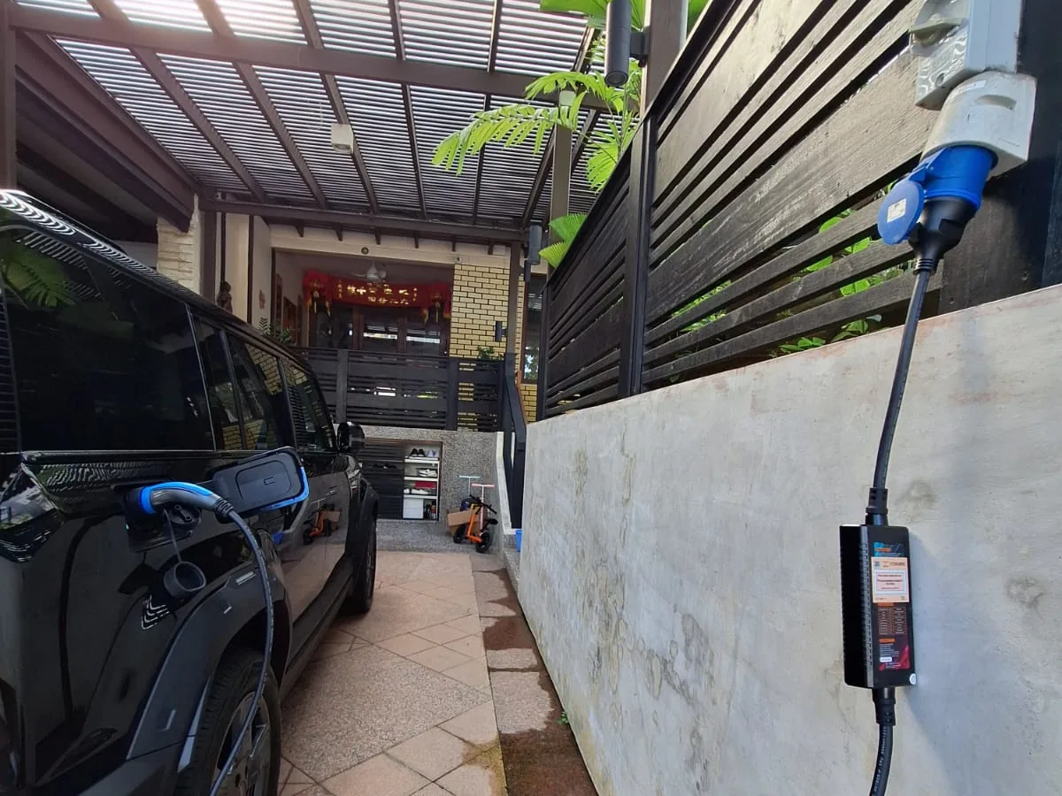 A dark SUV charging along a narrow covered side driveway from a blue industrial socket mounted high on the wall, with the portable control box hanging below it.