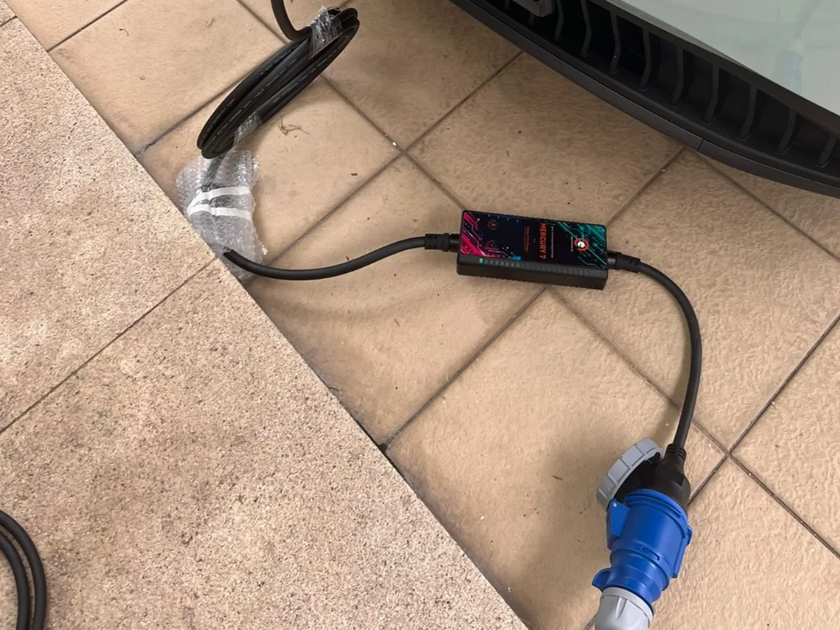 A portable EV charging set laid out on tiled floor — an in-line control box with a cable running to a blue industrial plug, beside a car's tyre.