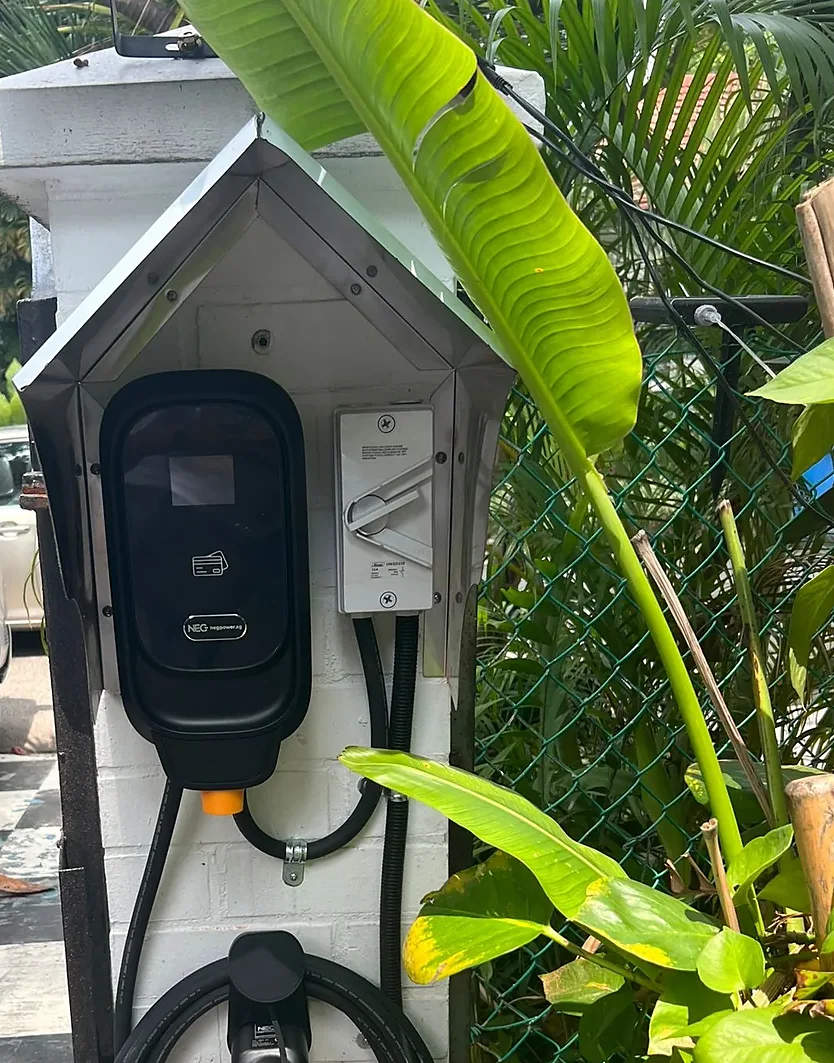 A stainless steel pitched canopy fixed over a wall-mounted EV charger and its isolator on a painted gate pillar, with planting and a wire fence beside it.