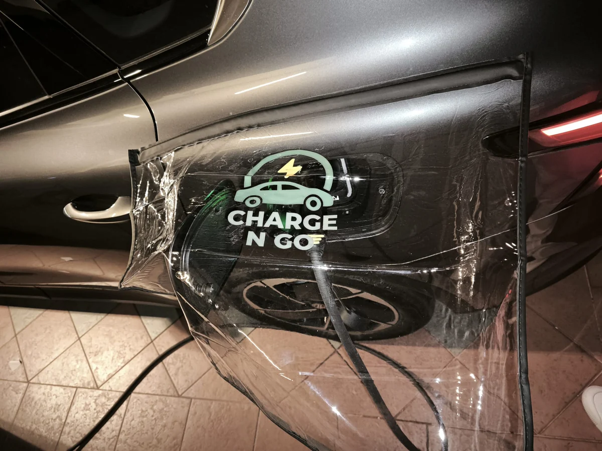 A clear Charge N Go branded rain shield draped over the charging port area of a car, the charging cable running underneath it.