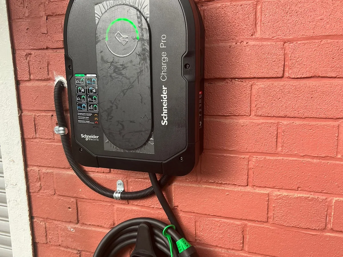 A black Schneider Charge Pro wall charger mounted on a red-painted brick wall, its green illuminated ring lit, flexible metal conduit entering from the left and the tethered cable coiled below.