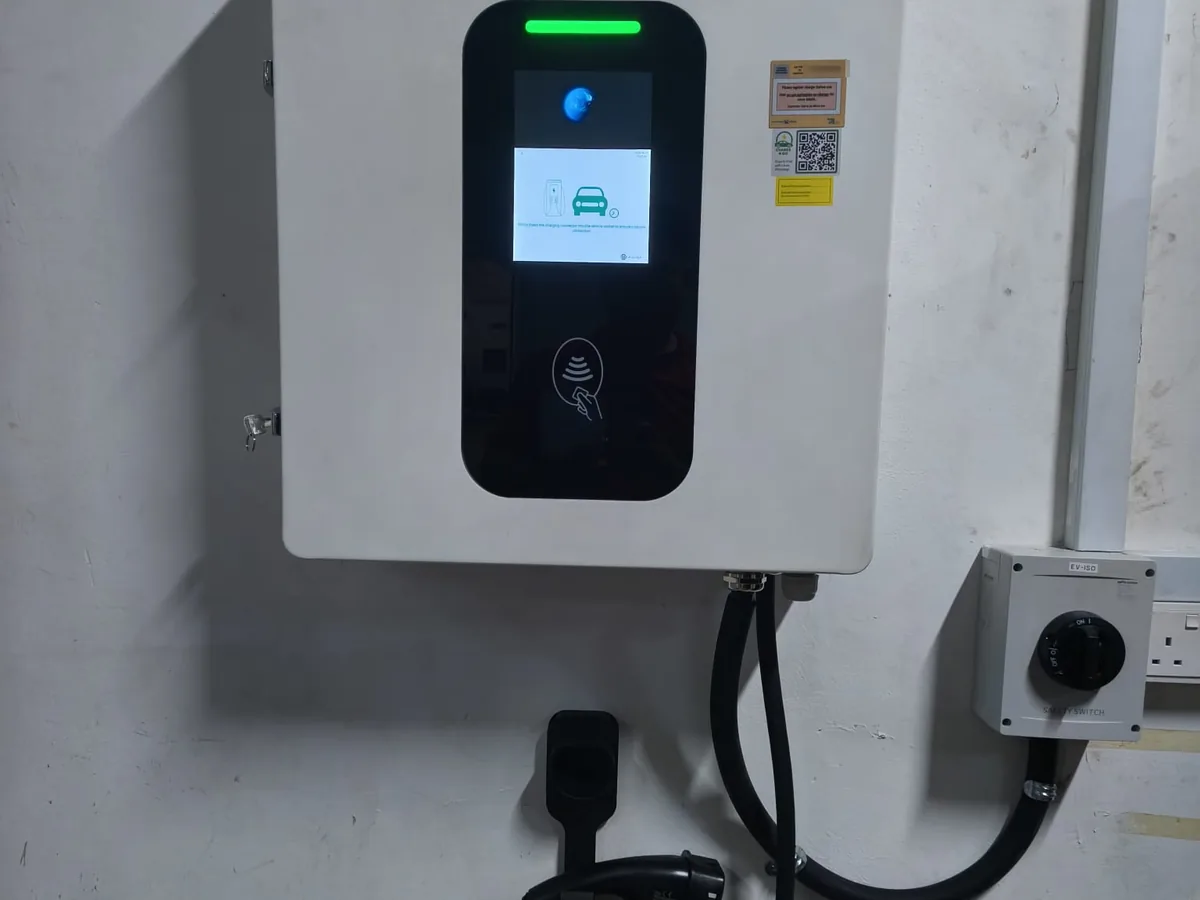 A wall-mounted EV charger with a colour display, a green status bar and an RFID reader, an orange LTA approval label above it and its own isolator switch on the wall alongside.