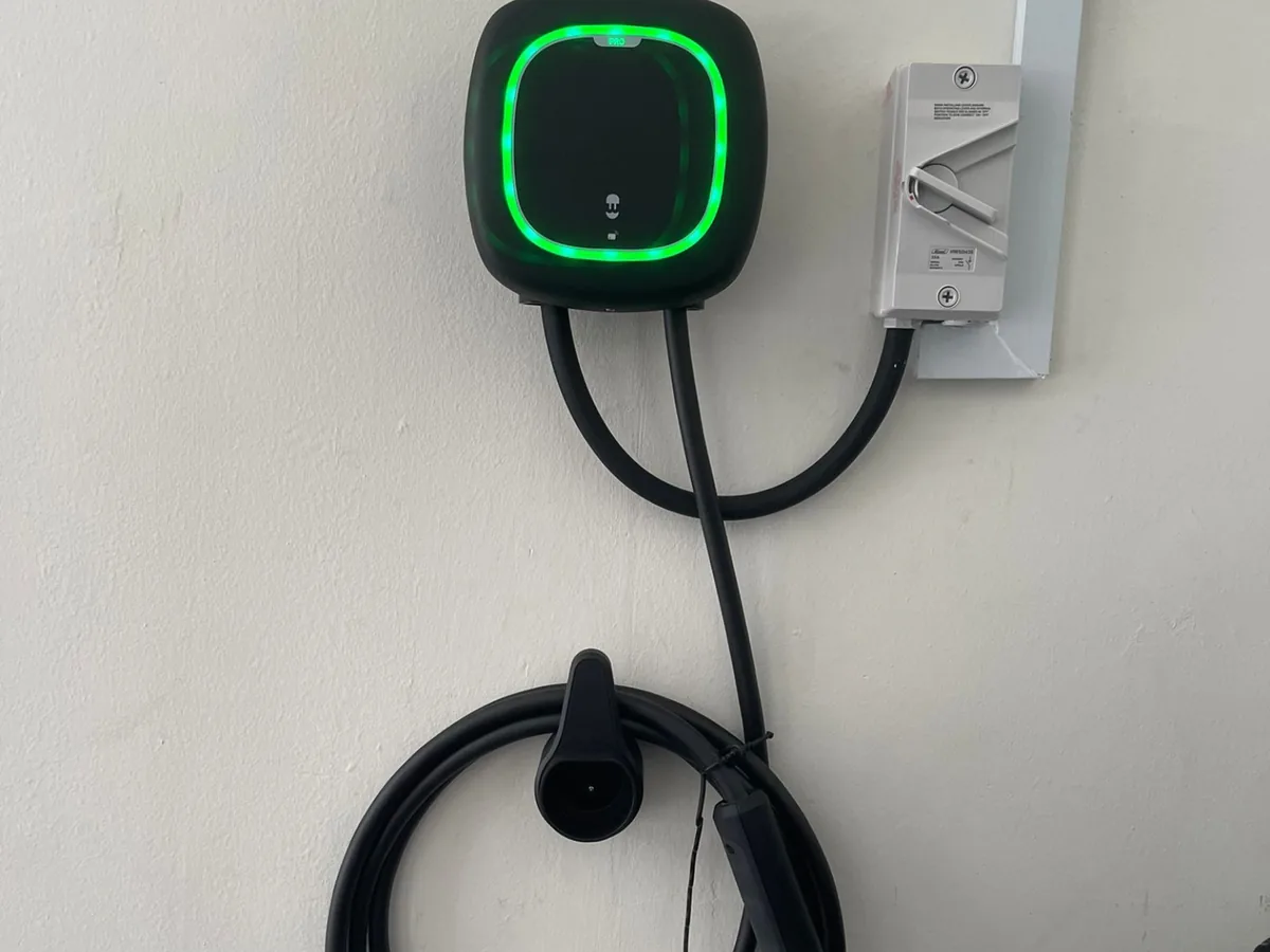 A round-faced wall charger with a bright green illuminated ring on a plain white wall, an isolator switch to the right and the cable hanging on a holster.