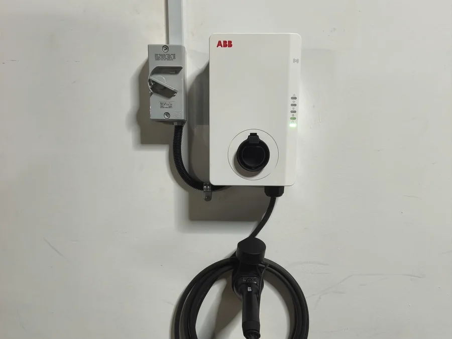 A white ABB wall charger with a black socket cap, mounted on a plain wall with an isolator above it and the cable coiled below.