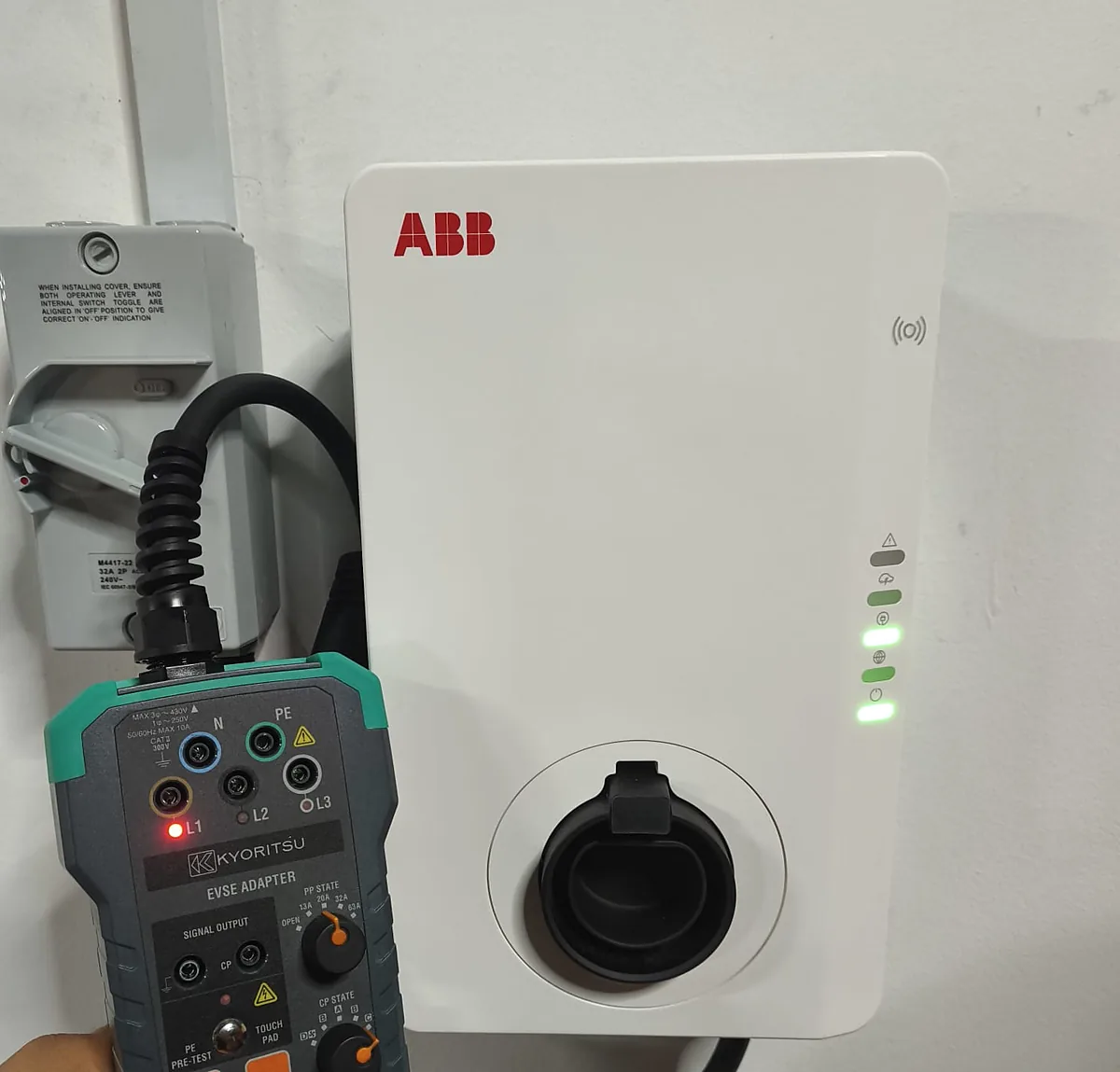 A white ABB wall charger, its status LEDs lit green, with a Kyoritsu test instrument held against it during commissioning.