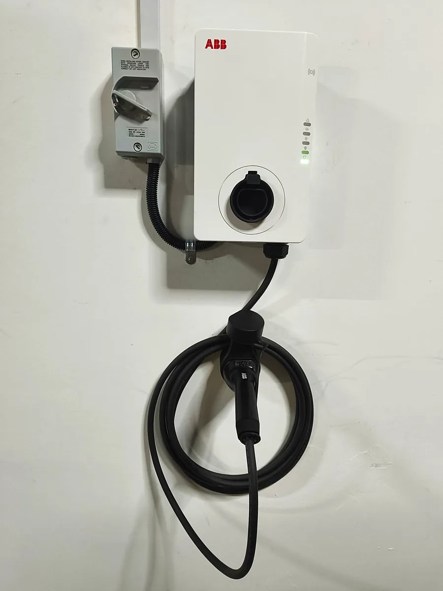 A white ABB wall charger mounted beside a switch enclosure, its tethered connector stowed on the holster below.