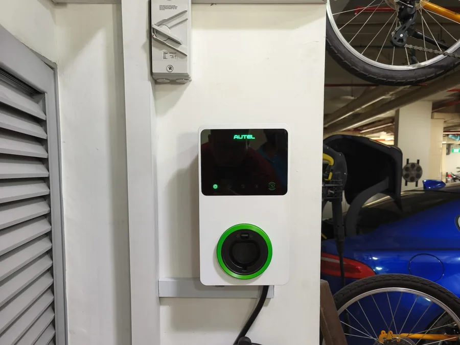 A white Autel wall charger with a green illuminated ring, mounted on a pillar in a covered storage area with bicycles behind.