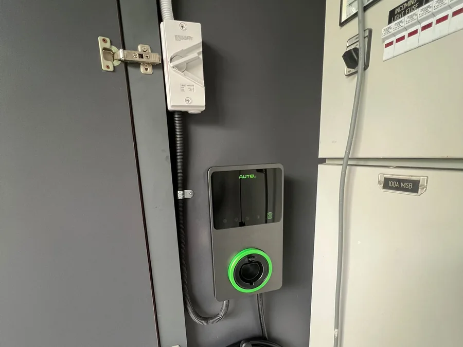 An Autel wall charger mounted beside service equipment and a distribution board, cable stowed on the holster below.