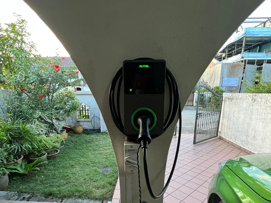 An Autel wall charger with a green ring mounted on a rendered outdoor pillar, a green car parked alongside and a garden behind.