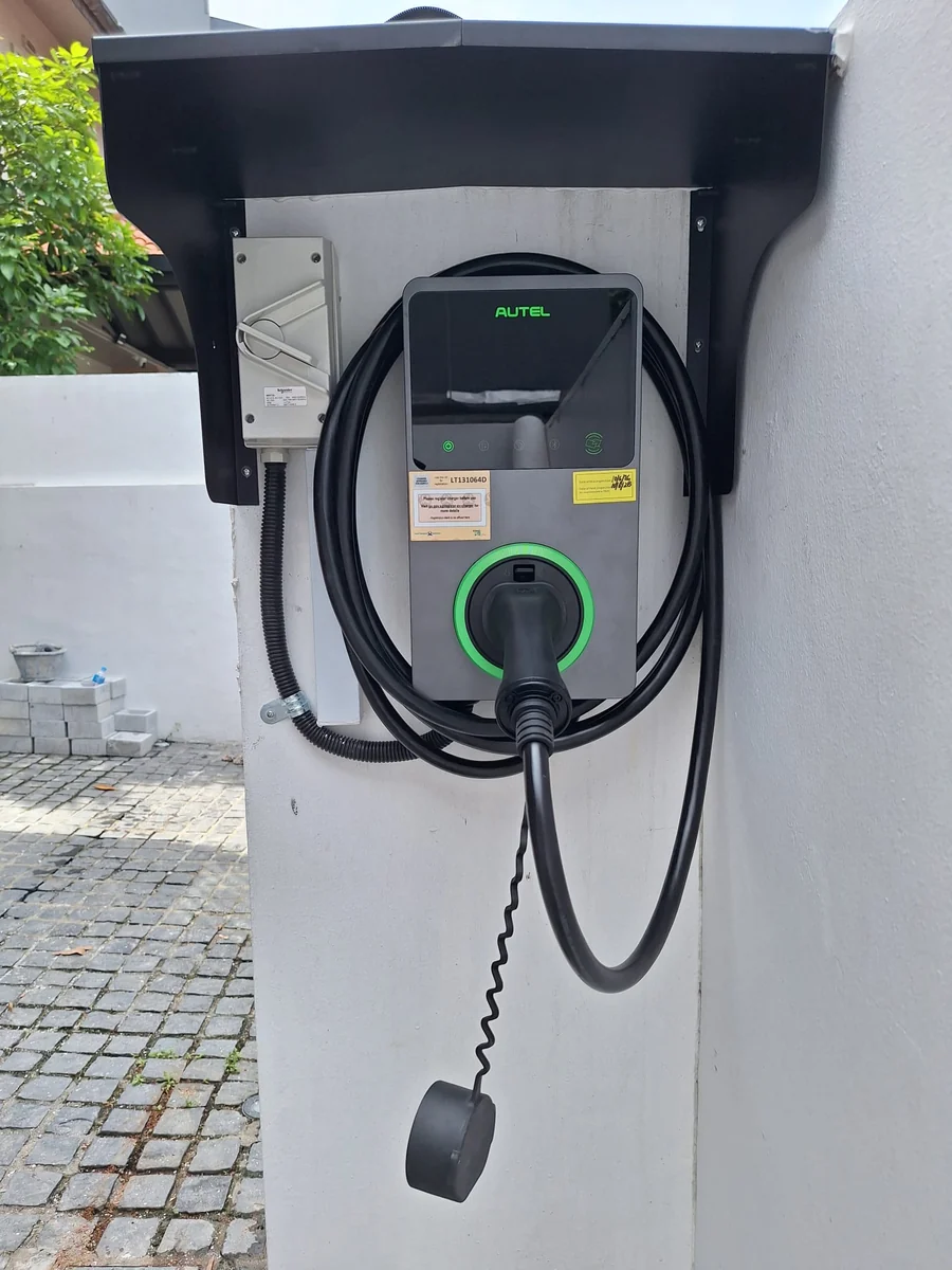 A dark Autel MaxiCharger with its ring lit green, mounted on a wall under a black steel weather hood with an isolator to the left and the connector on a holster.