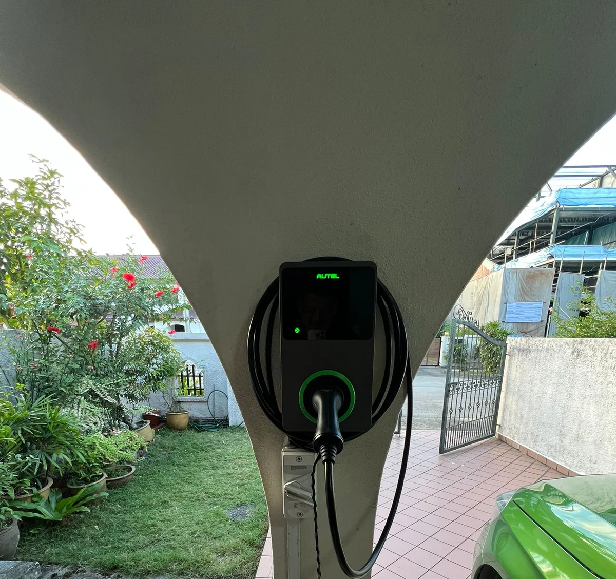 A dark Autel MaxiCharger on a tapered white pillar beside a lawn and flowering shrubs, its cable running down to a car parked on the tiled forecourt.