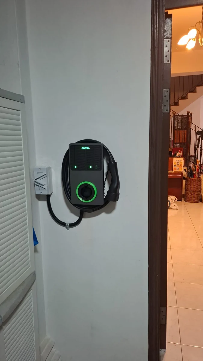 A dark Autel MaxiCharger with its ring lit green on a wall beside an open doorway, warm interior light and a tiled floor beyond.