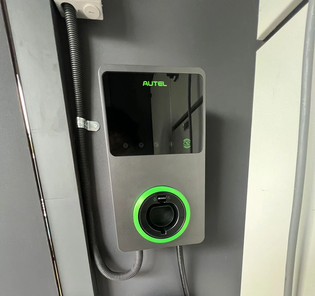 Close view of a grey Autel MaxiCharger showing the green wordmark, the touch icons and the illuminated green socket ring, with flexible conduit alongside.