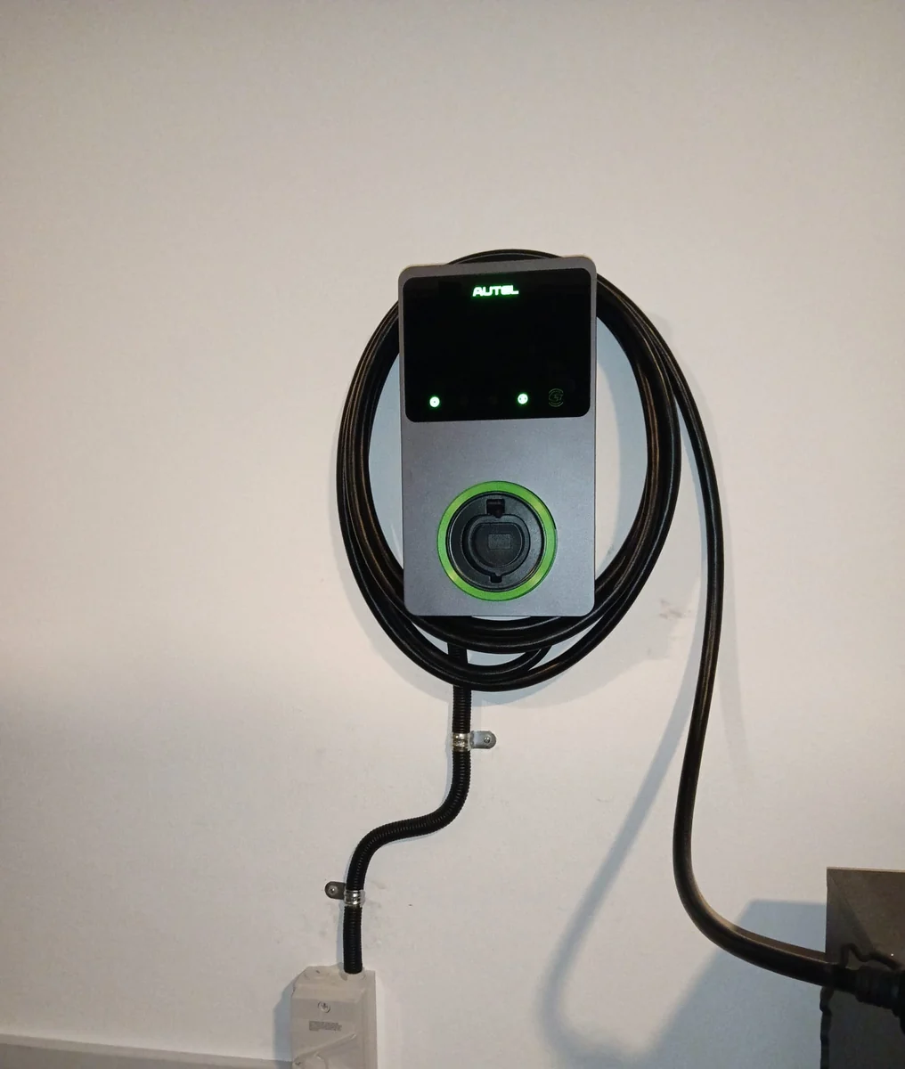 A grey Autel MaxiCharger on a plain wall with two status lights lit green and the tethered cable coiled in a wide loop beneath it.