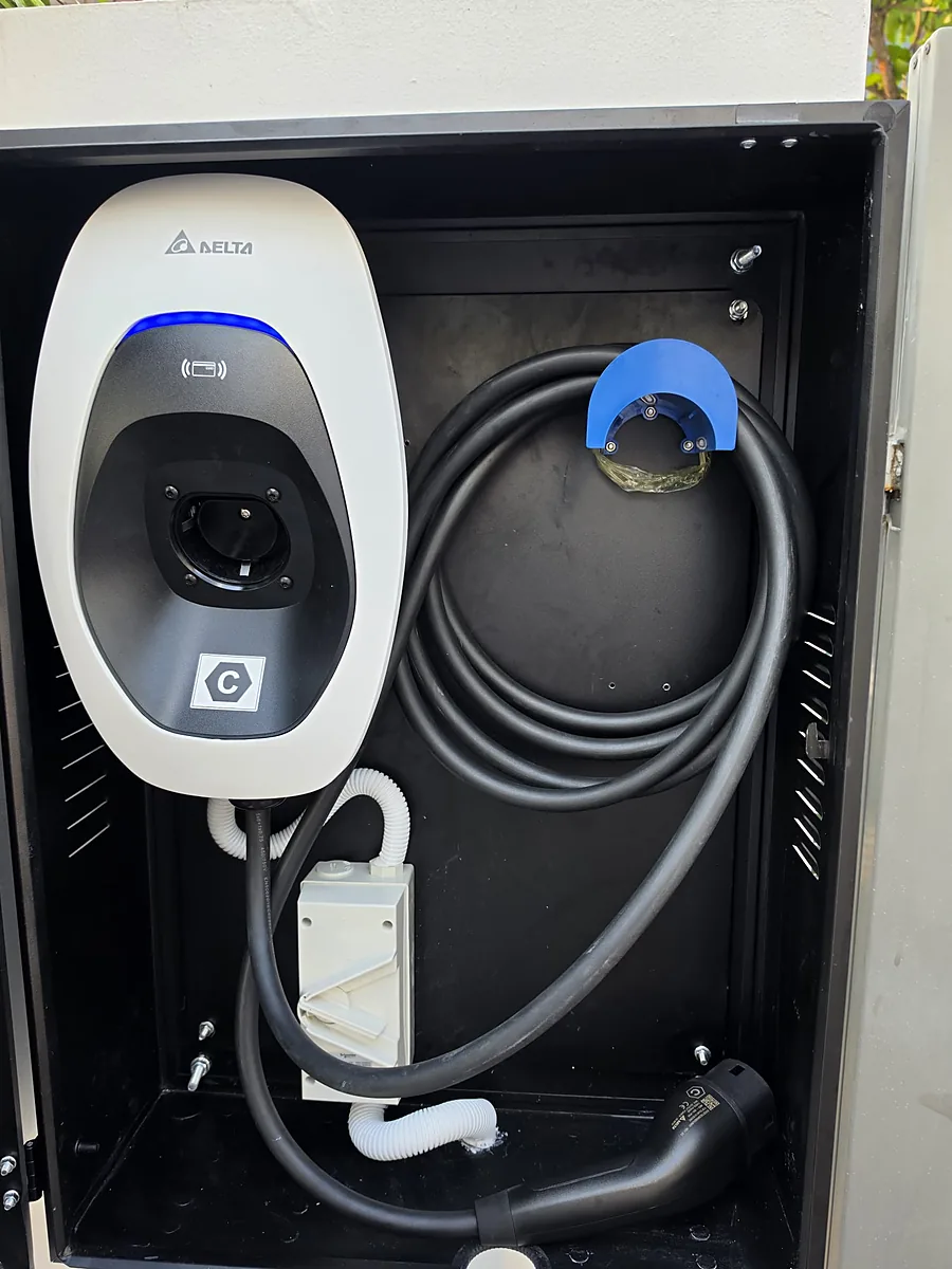 A white Delta EV charger with a blue status ring mounted inside an open black weatherproof cabinet, its charging cable coiled on a hanger beside it and white flexible conduit running down to a Schneider weatherproof isolator.