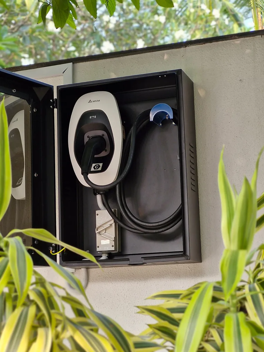 An open black weatherproof cabinet on a rendered exterior wall housing a white Delta EV charger, its connector plugged into the unit's socket and the cable coiled behind a blue plastic hook, with the supply running in white flexible conduit to a metal-clad isolator switch below.