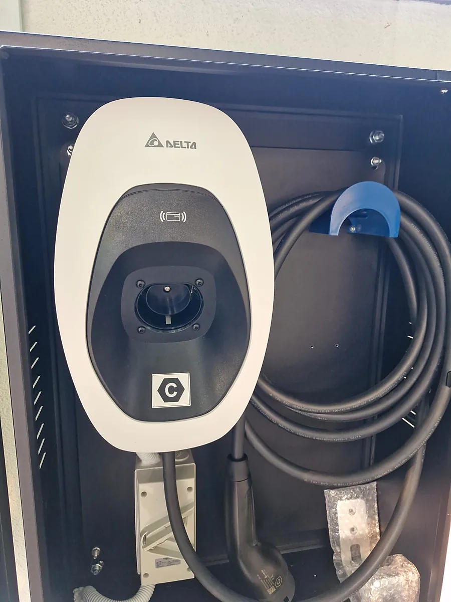 A white Delta EV charger with its outlet cover closed, mounted inside a black steel cabinet, a charging cable coiled on the back panel beside a blue holster and a white weatherproof enclosure on flexible conduit below the unit.