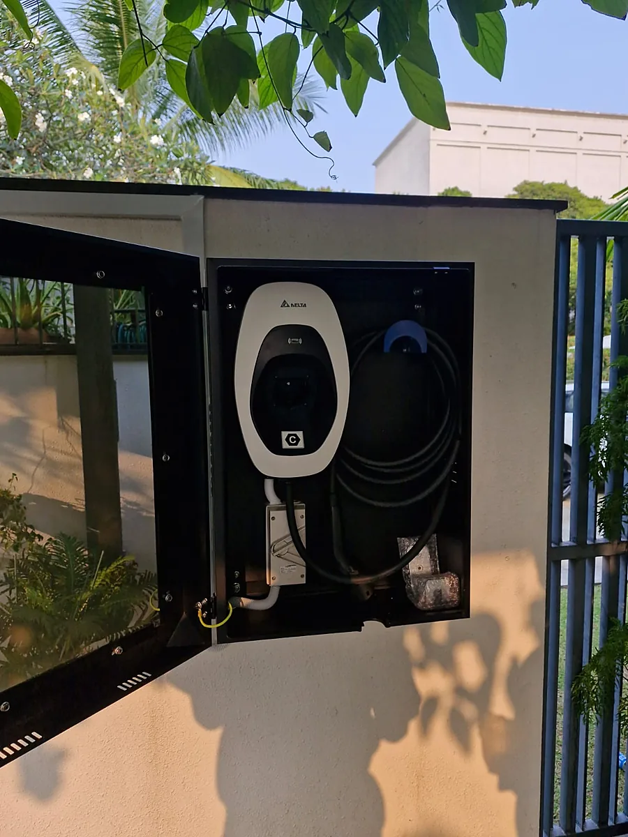 An open black weatherproof cabinet on an exterior rendered pillar housing a white Delta EV charger, its coiled cable on a blue holster and a grey metal enclosure on white flexible conduit below, with a garden railing and planting beyond.