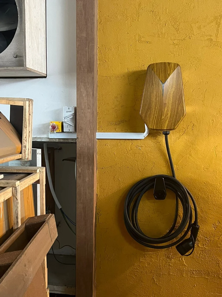 A wood-grain HiCi charger on a mustard-yellow wall in a workshop, timber crates stacked to the left and the cable coiled on its holster.
