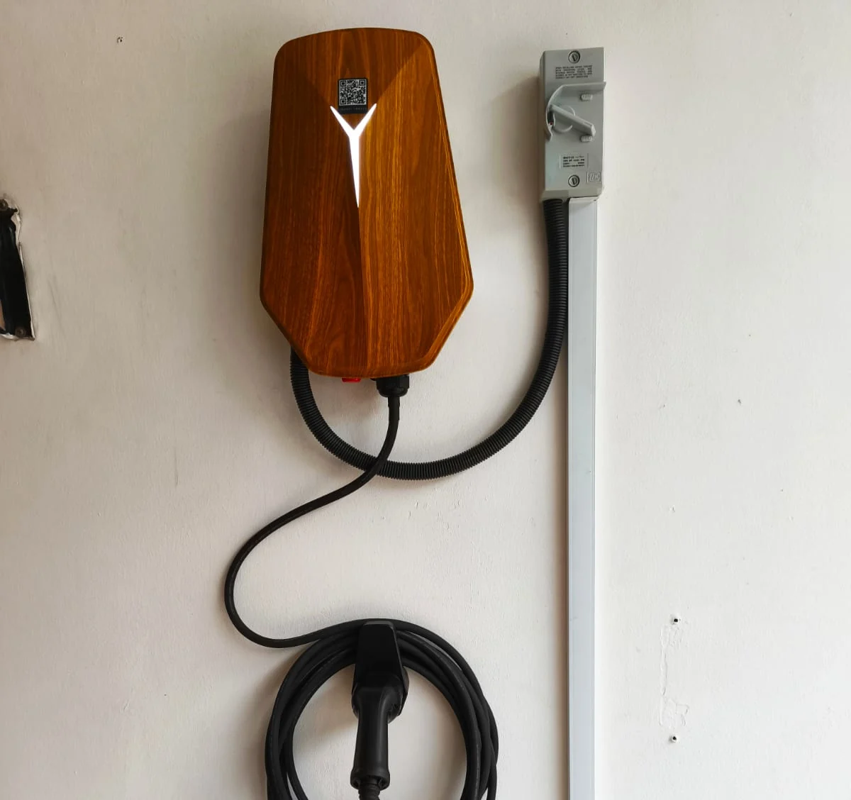 A wood-grain HiCi charger with a QR label on a pale wall, a grey isolator switch mounted to its right and the cable curving down to a holster.