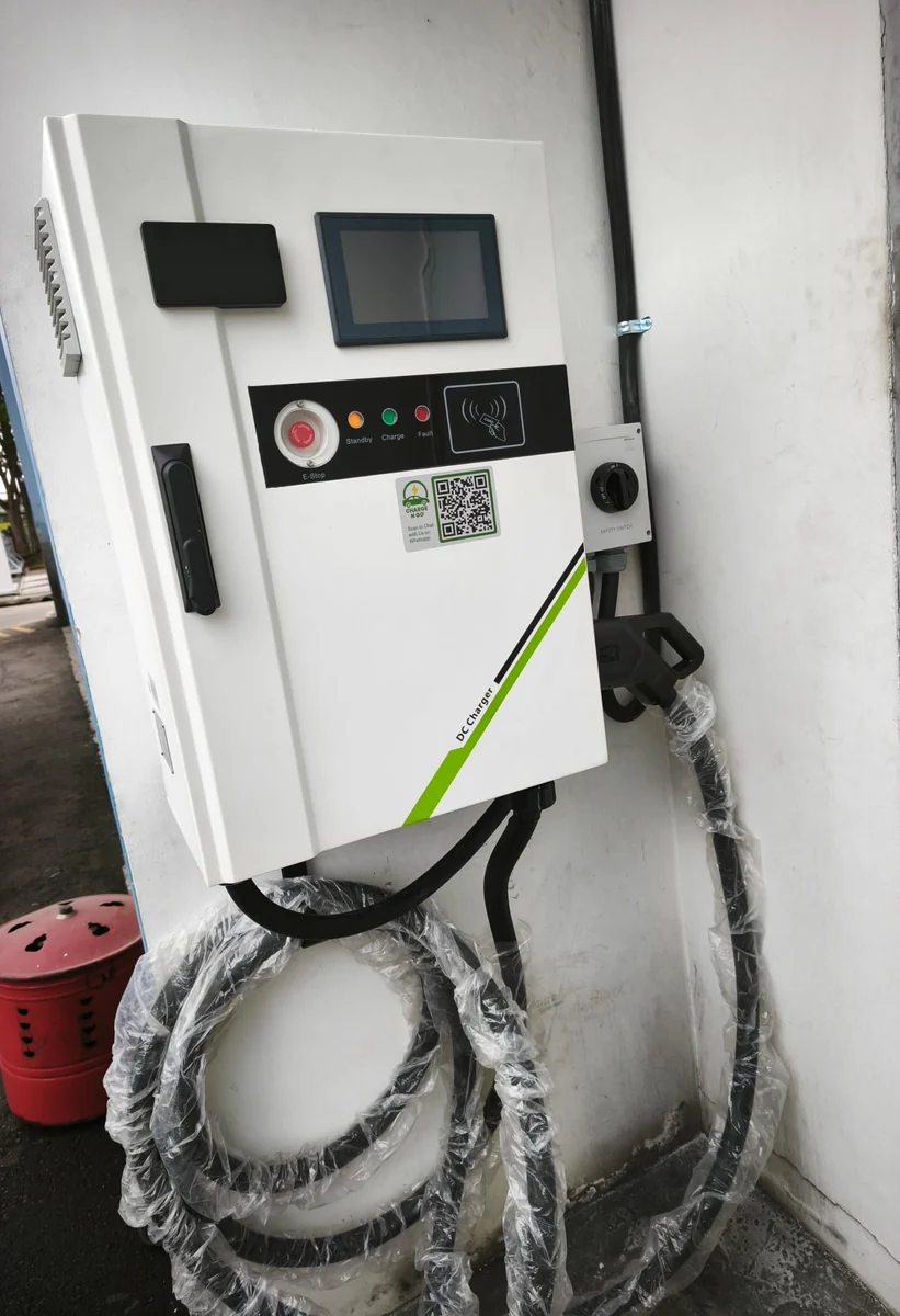 A white wall-mounted DC charger seen at an angle, showing the touchscreen, status lights, emergency stop and a green livery stripe, its cables wrapped below.