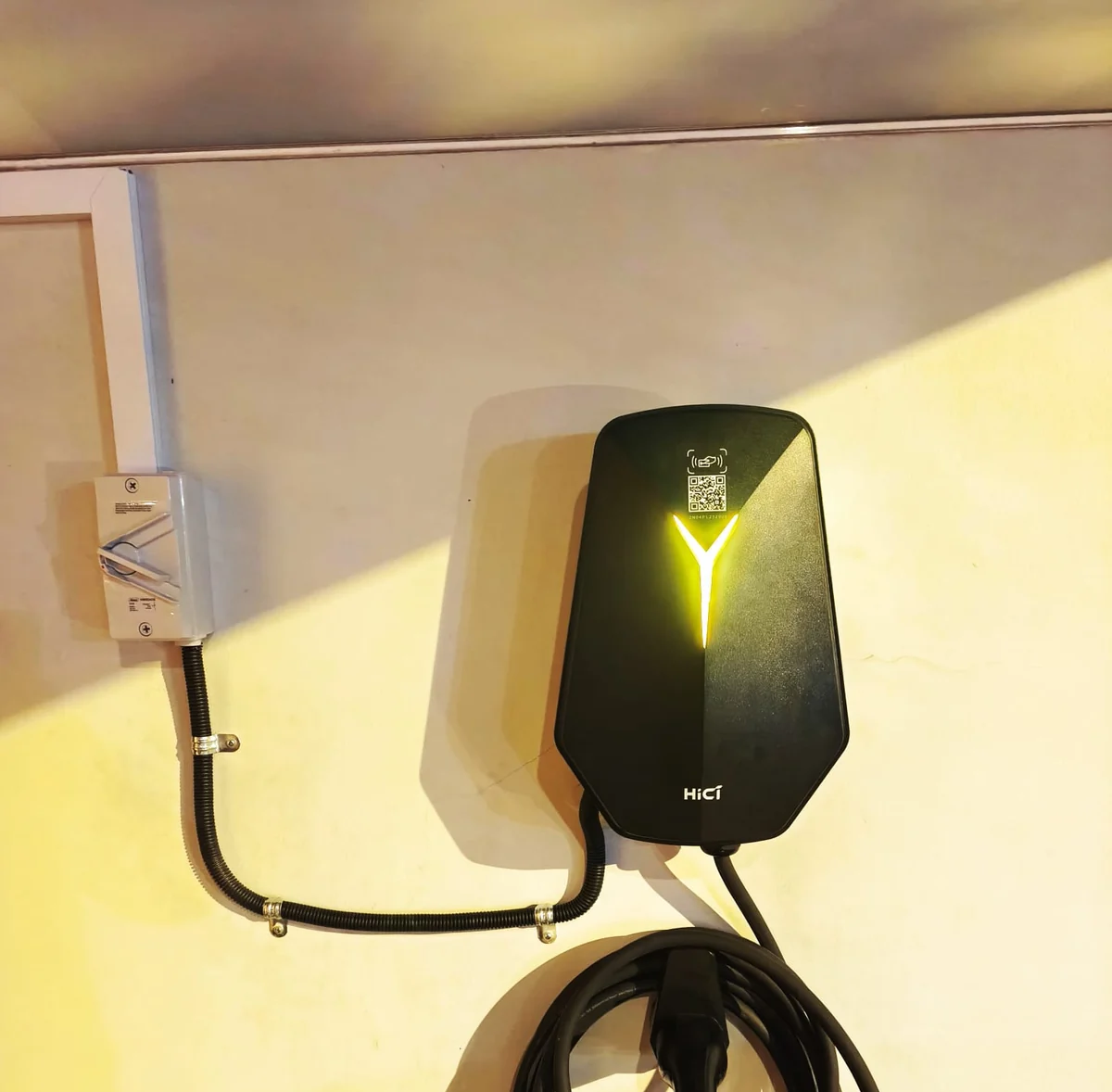 A black faceted HiCi charger with its Y light lit, mounted under a canopy in warm evening light with black conduit entering from the left.