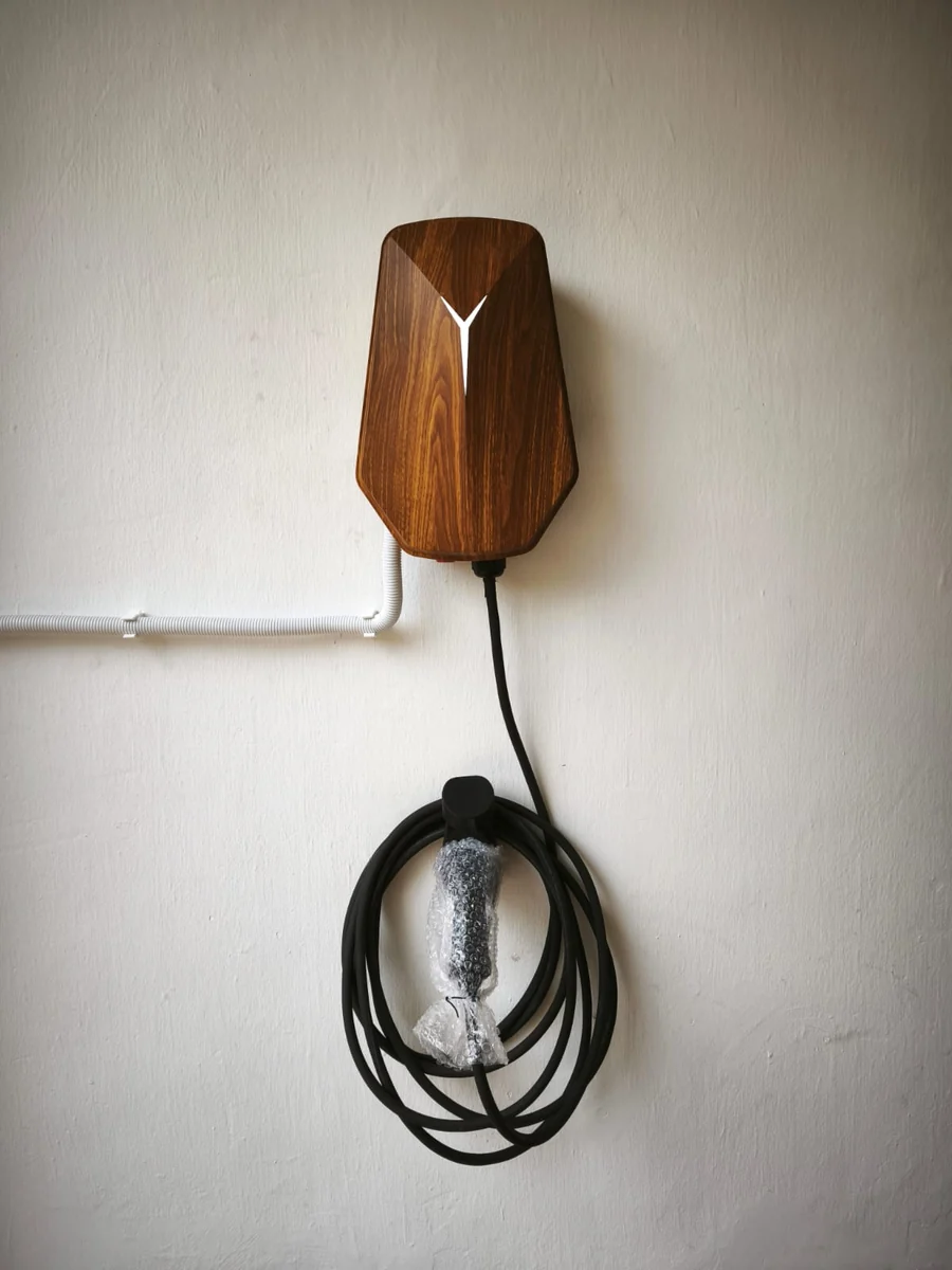 A wood-grain HiCi charger on a plain wall with white conduit entering horizontally and the cable coiled beneath in a protective wrap.