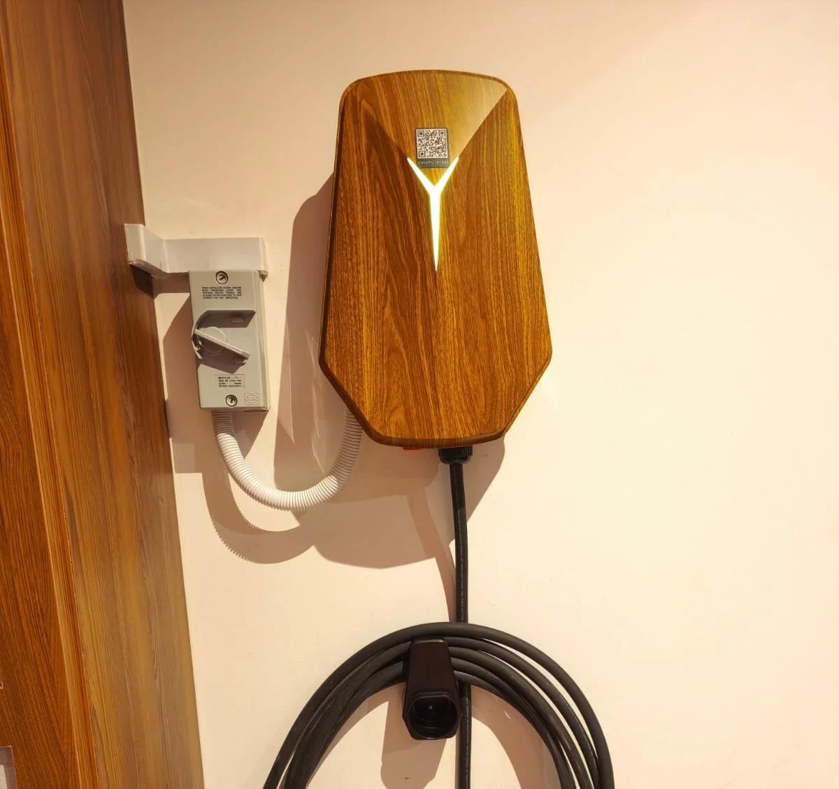 A wood-grain HiCi charger on a pale wall next to a timber door frame, with an isolator and white conduit to its left and the cable coiled below.