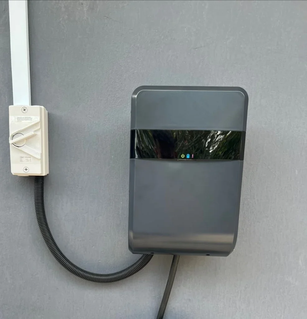 A grey Lite-On wall charger on a grey rendered wall with a cream isolator switch to its left, flexible conduit looping between the two and greenery reflected in the glossy status band.