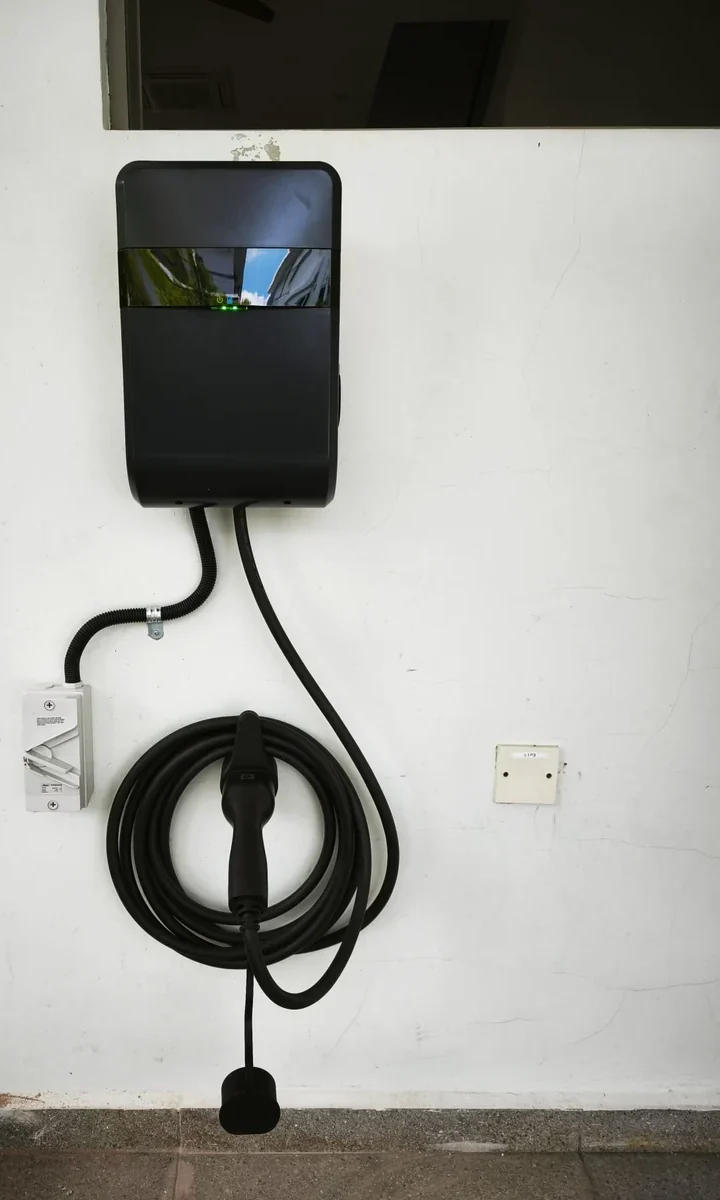 A dark grey Lite-On wall charger on a white wall below a dark window opening, its tethered cable coiled on a stainless holster bracket with the connector stowed, and a wall socket to the right.