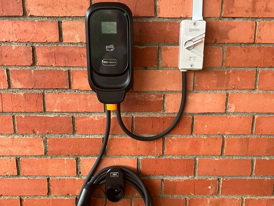 A black NEG wall charger mounted on a red brick wall with a white isolator switch beside it and the tethered connector hanging below.