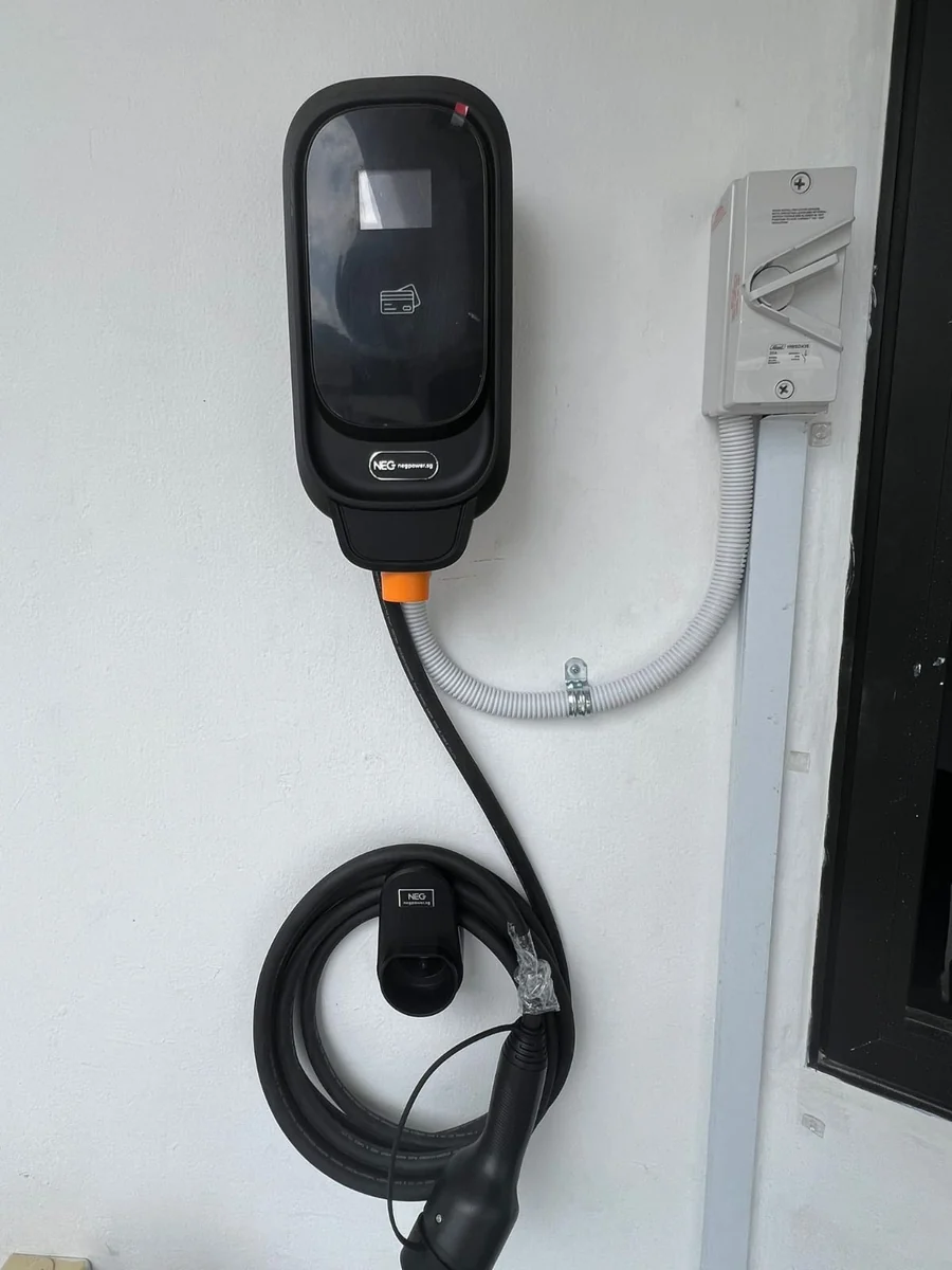A black NEG charger on a plain white wall with an isolator to the right, the cable dropping to a coiled loop and connector.