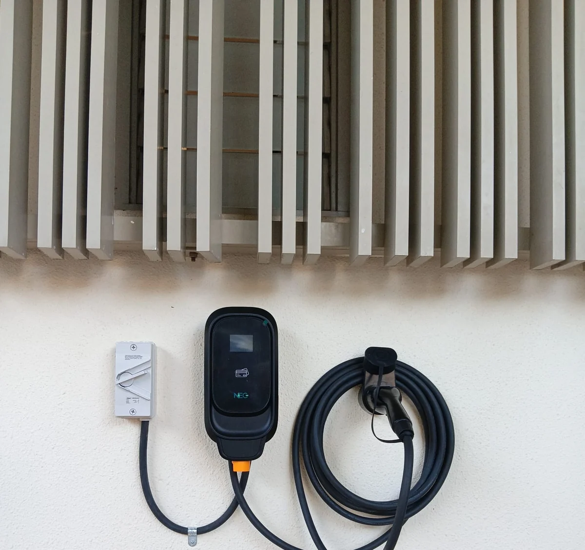 A black NEG charger on a white wall beneath timber louvre screening, an isolator to its left and the cable coiled alongside.