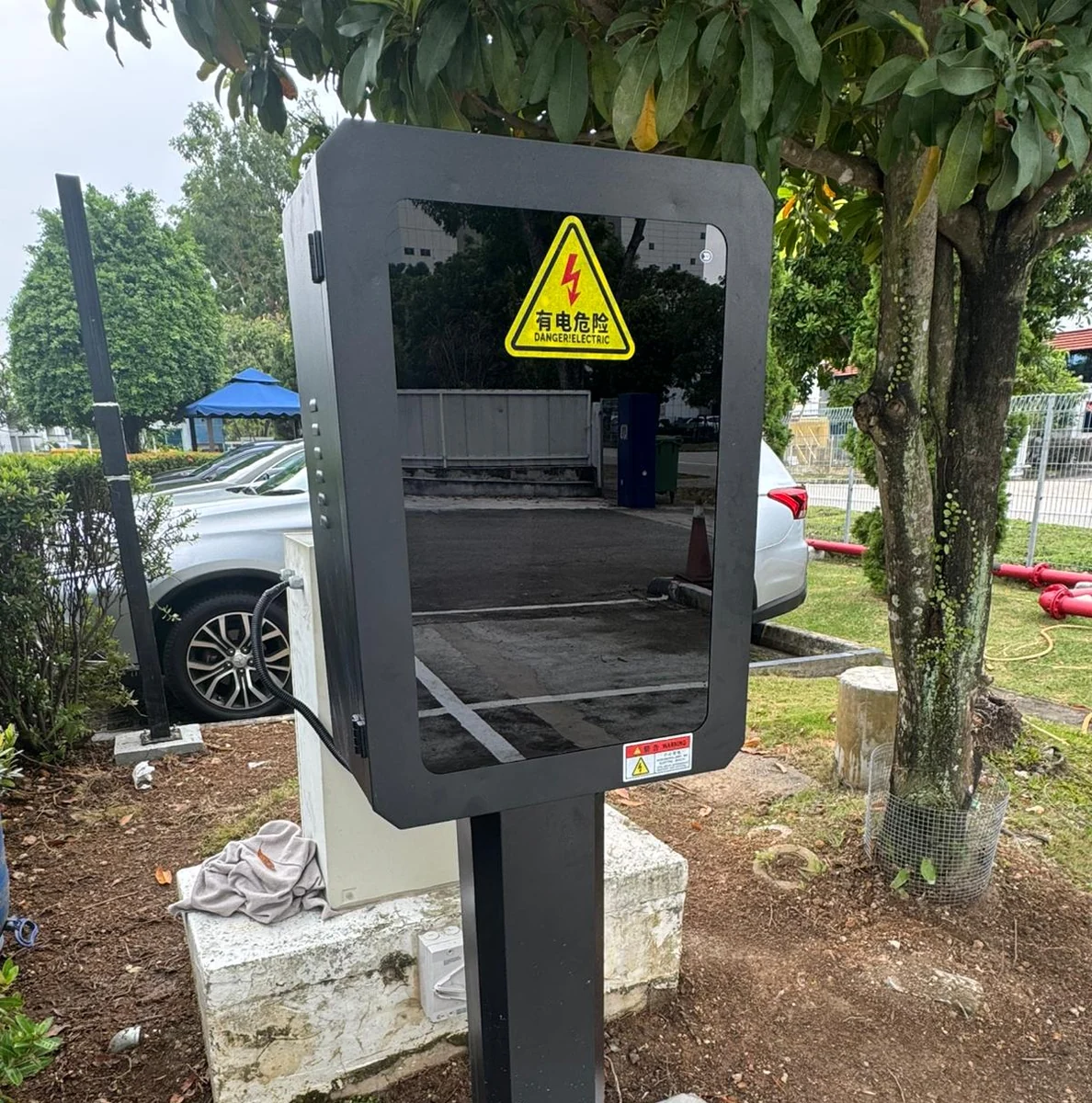 A charger inside a black post-mounted enclosure with a warning label on the glazed door, standing in a planted bed beneath a mature tree.