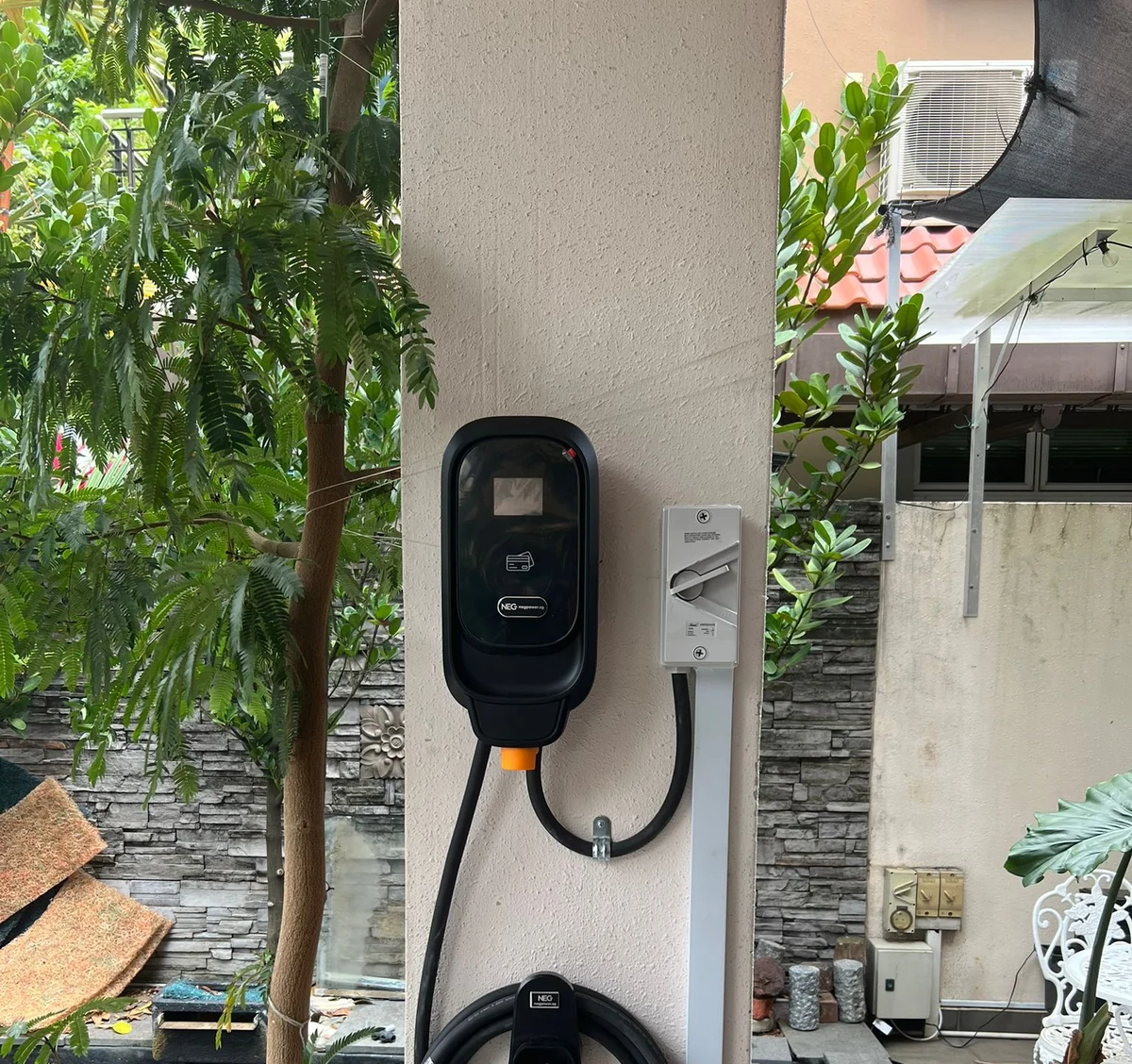 A black NEG charger on a cream wall in a landed-home garden, palms and shrubs either side and the connector stowed below.