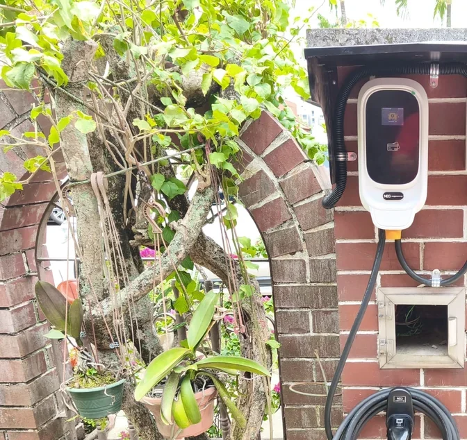 A white NEG charger under a stone coping on a red brick archway, with orchids and a mature climbing tree either side and the connector on its holster.