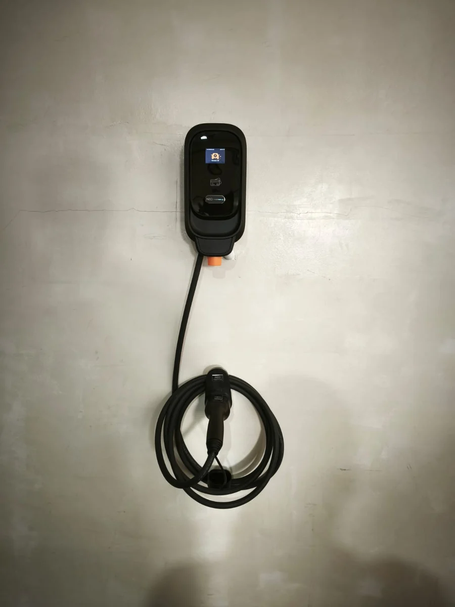 A black NEG charger on a plain rendered wall, cable coiled neatly beneath it.