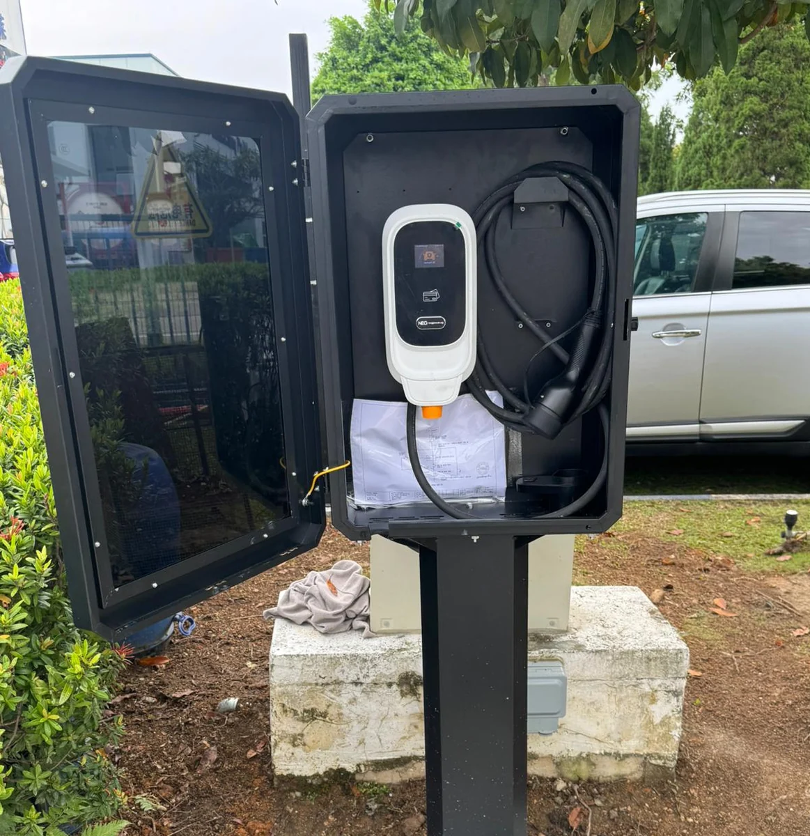 A black post-mounted enclosure standing open on a concrete plinth, a white NEG charger and its coiled cable inside, lawn behind.